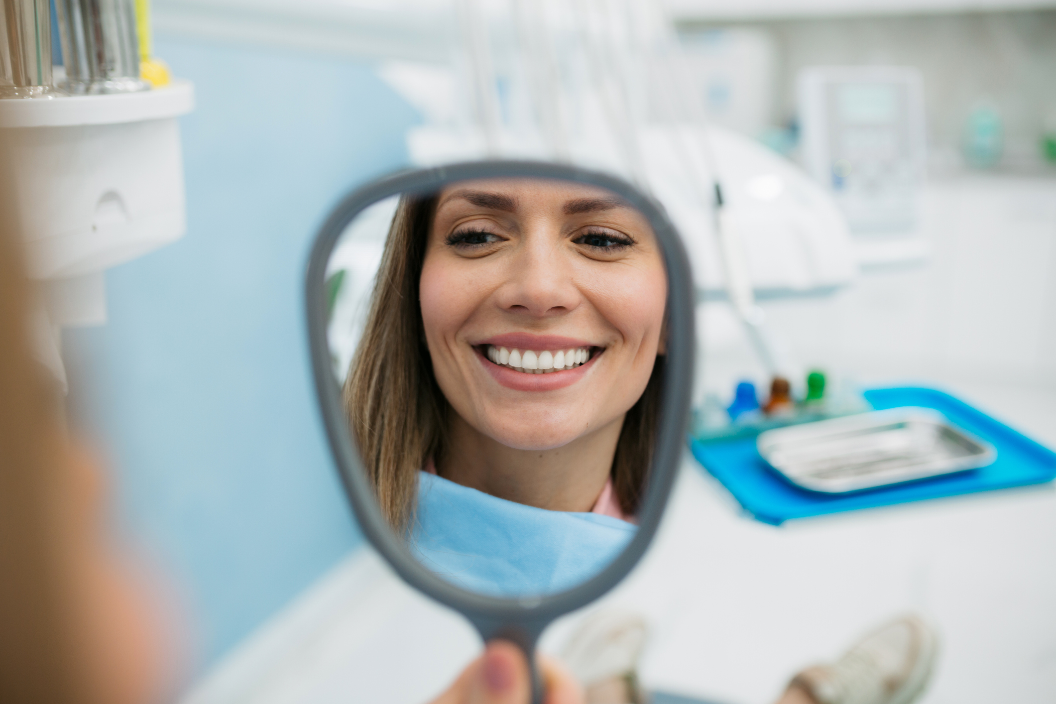 Young adult happy female patient relaxing while visiting a dentist office, holding a vanity mirror and with a toothy smile on her face inspecting her teeth