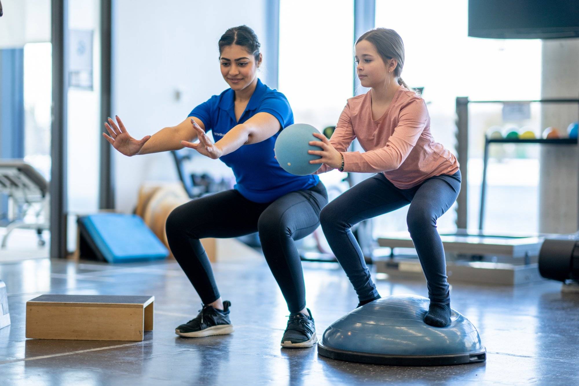 A young girl works with her physiotherapist in a gym during an appointment.  She is dressed comfortably and is balancing on a half ball as she follows the therapist instructions and does a squat with a ball in her hands.